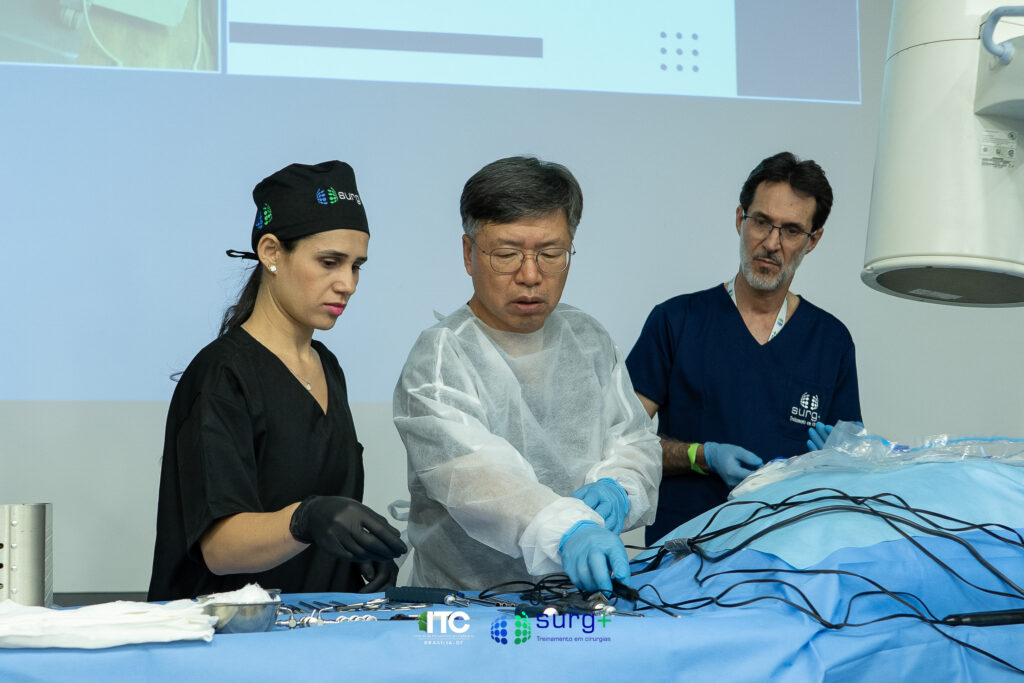 À esquerda da imagem, você vê uma instrumentadora. No centro está o criador da Endoscopia Biportal, Dr. Sangkyu Son. À direita da imagem, auxiliando o Dr. Son, está o Dr. Eidmar Neri — um dos pioneiros da técnica no Brasil. Os três estão organizando os instrumentos cirúrgicos e os outros preparativos para realizar a demonstração da UBE na prática.